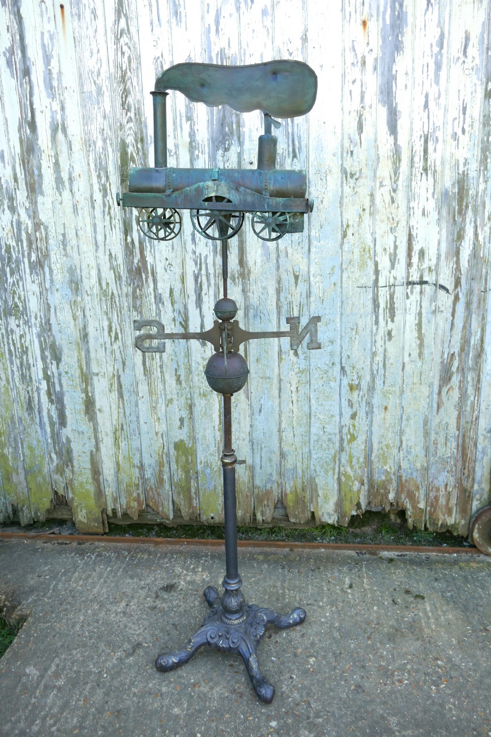 A nineteenth-century copper weather vane shaped like a steam locomotive train with verdigris patina, featuring directional cardinal points on a cast iron stand.