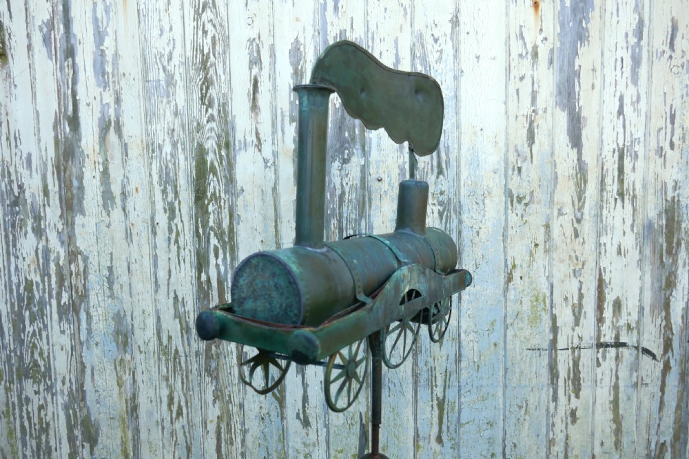 A nineteenth-century copper weather vane shaped like a steam locomotive train with verdigris patina, featuring directional cardinal points on a cast iron stand. View III