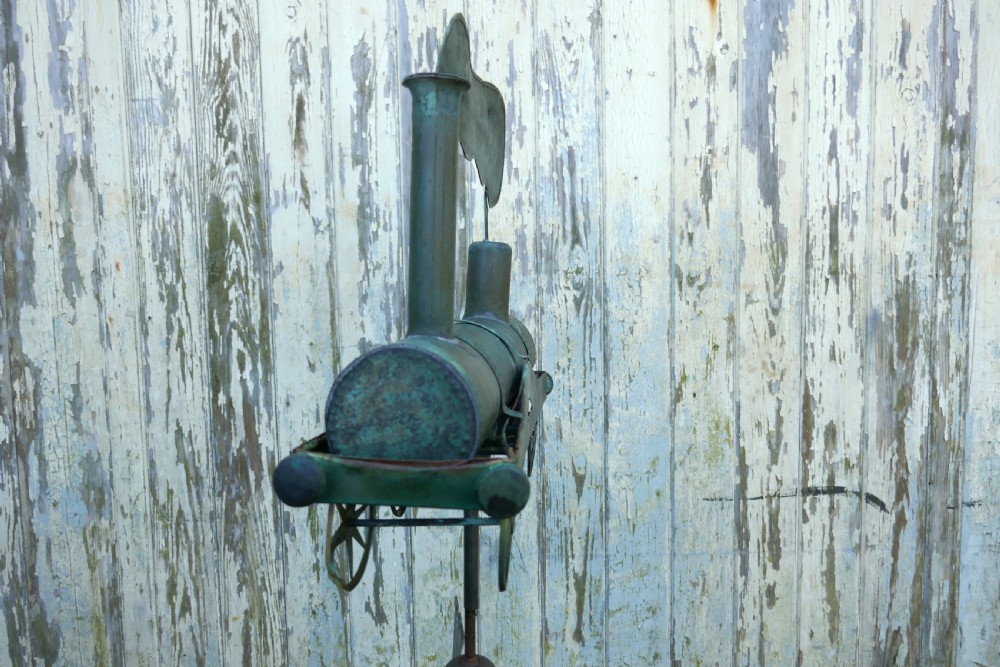 A nineteenth-century copper weather vane shaped like a steam locomotive train with verdigris patina, featuring directional cardinal points on a cast iron stand. View IV
