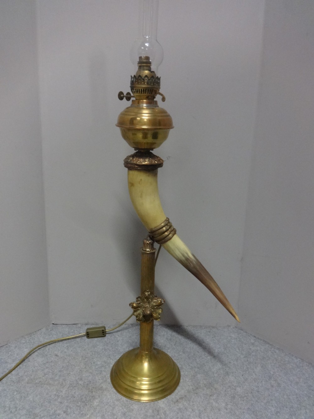 A Victorian table lamp featuring a curved natural horn stem mounted on a fluted brass pedestal base with a brass oil reservoir and clear glass chimney.