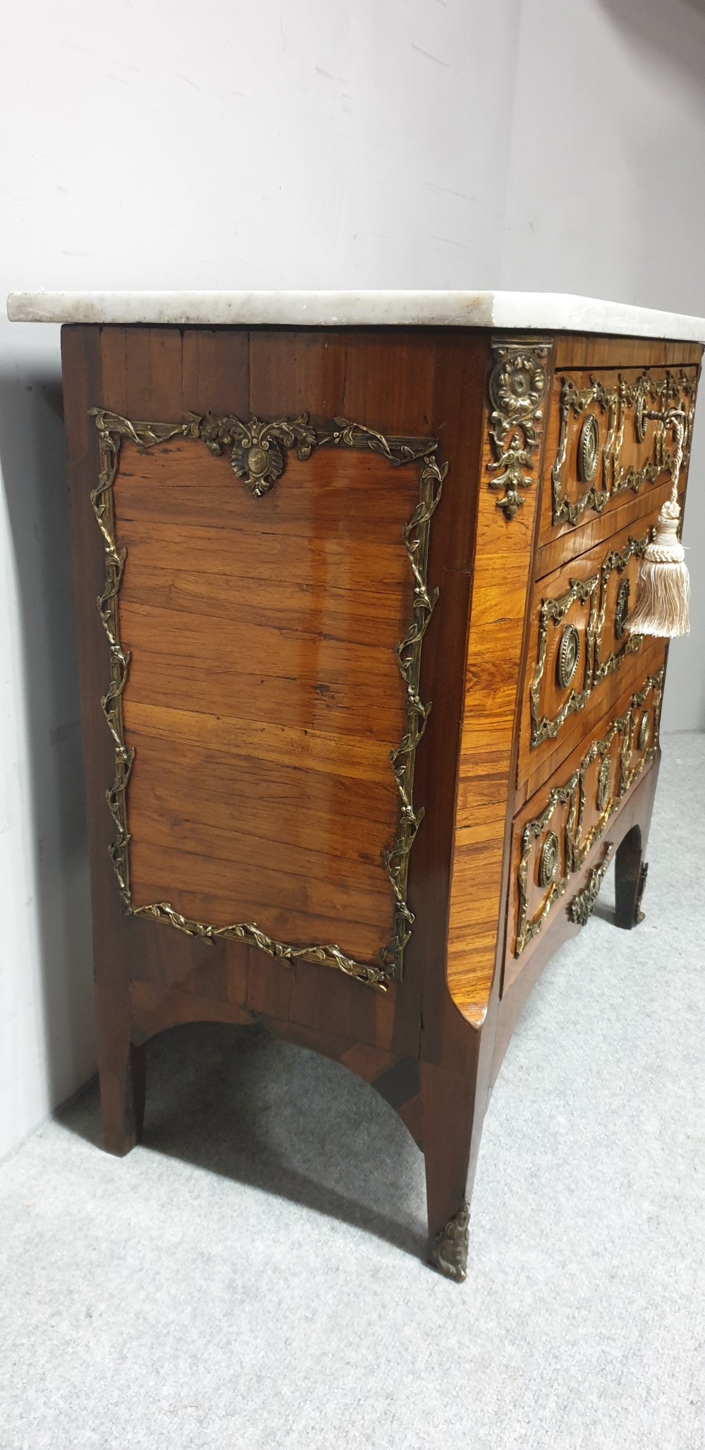 An eighteenth-century French commode chest of drawers featuring a white marble top, patterned wood veneer, and decorative gilt-bronze drawer mounts. View V
