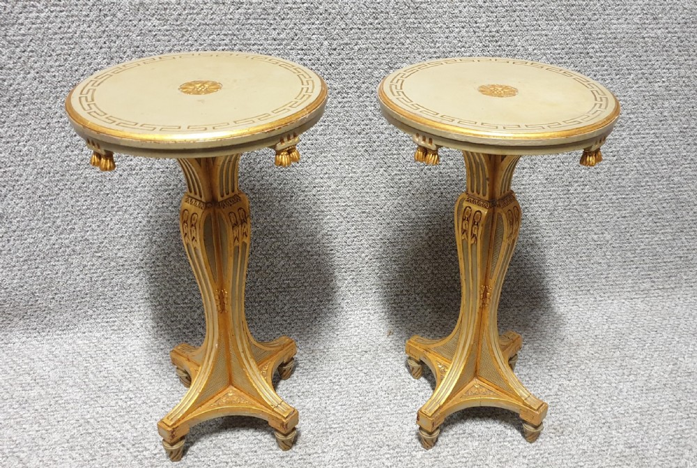 A pair of round painted side tables featuring cream tops with Greek key borders, gilded tassel details, and carved quadform pedestal bases.