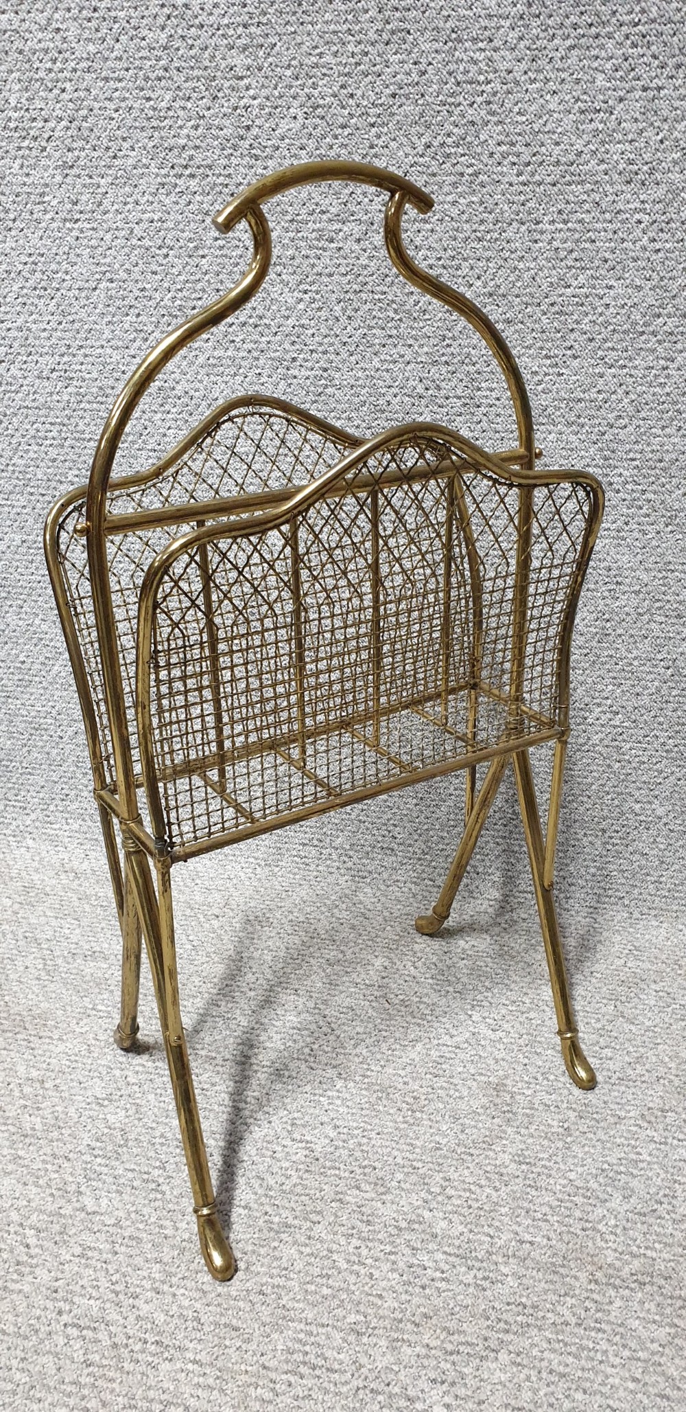 An Edwardian brass magazine rack featuring a tubular carrying handle, wire mesh sides with a central divider, and splayed legs.