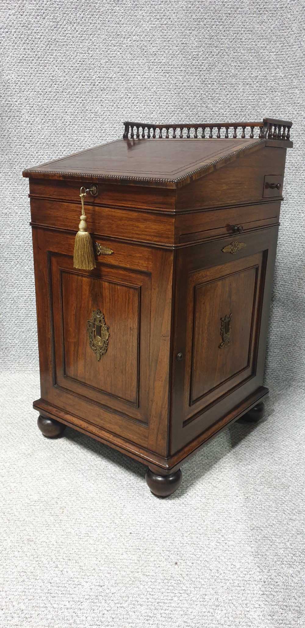 A Regency rosewood Davenport desk with a slanted leather writing slope, brass decorative mount, side drawers, and turned bun feet. View X