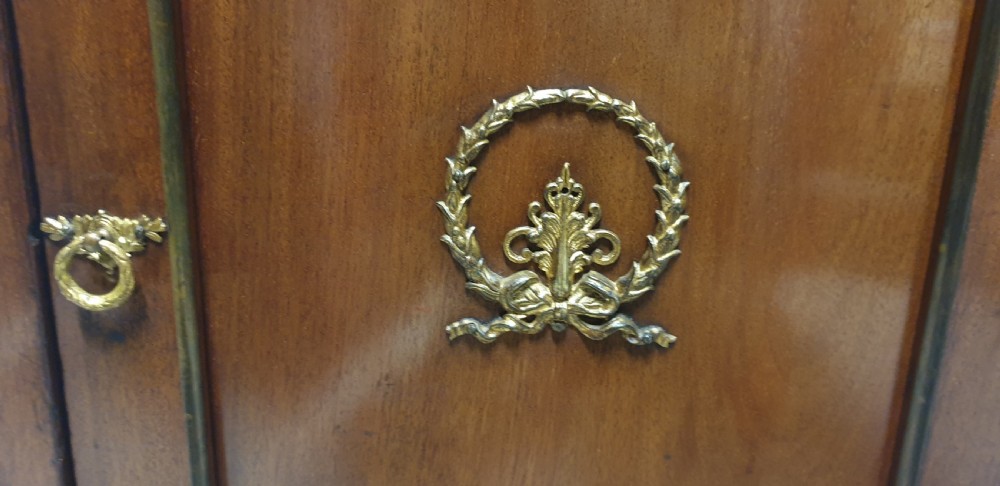 A mahogany side cabinet featuring gilt bronze mounts, a single top drawer over a cupboard door with a central laurel wreath, raised on square tapered legs. View IV