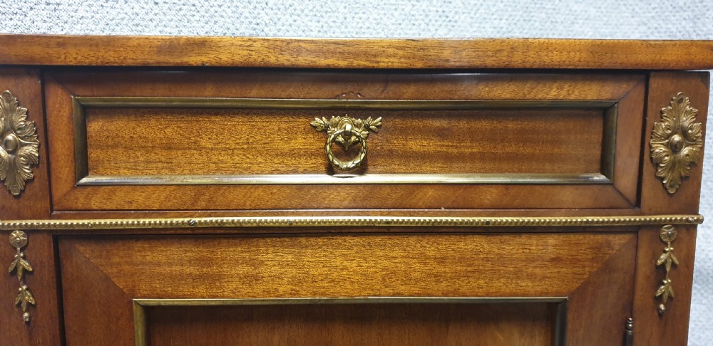 A mahogany side cabinet featuring gilt bronze mounts, a single top drawer over a cupboard door with a central laurel wreath, raised on square tapered legs. View V