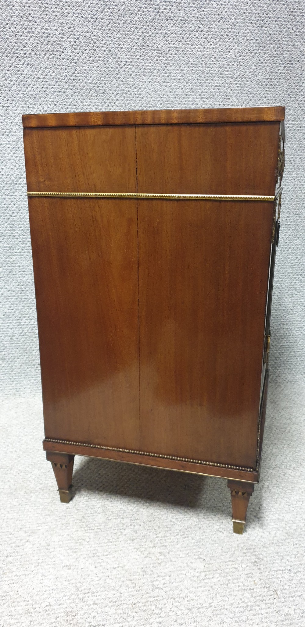 A mahogany side cabinet featuring gilt bronze mounts, a single top drawer over a cupboard door with a central laurel wreath, raised on square tapered legs. View VIII