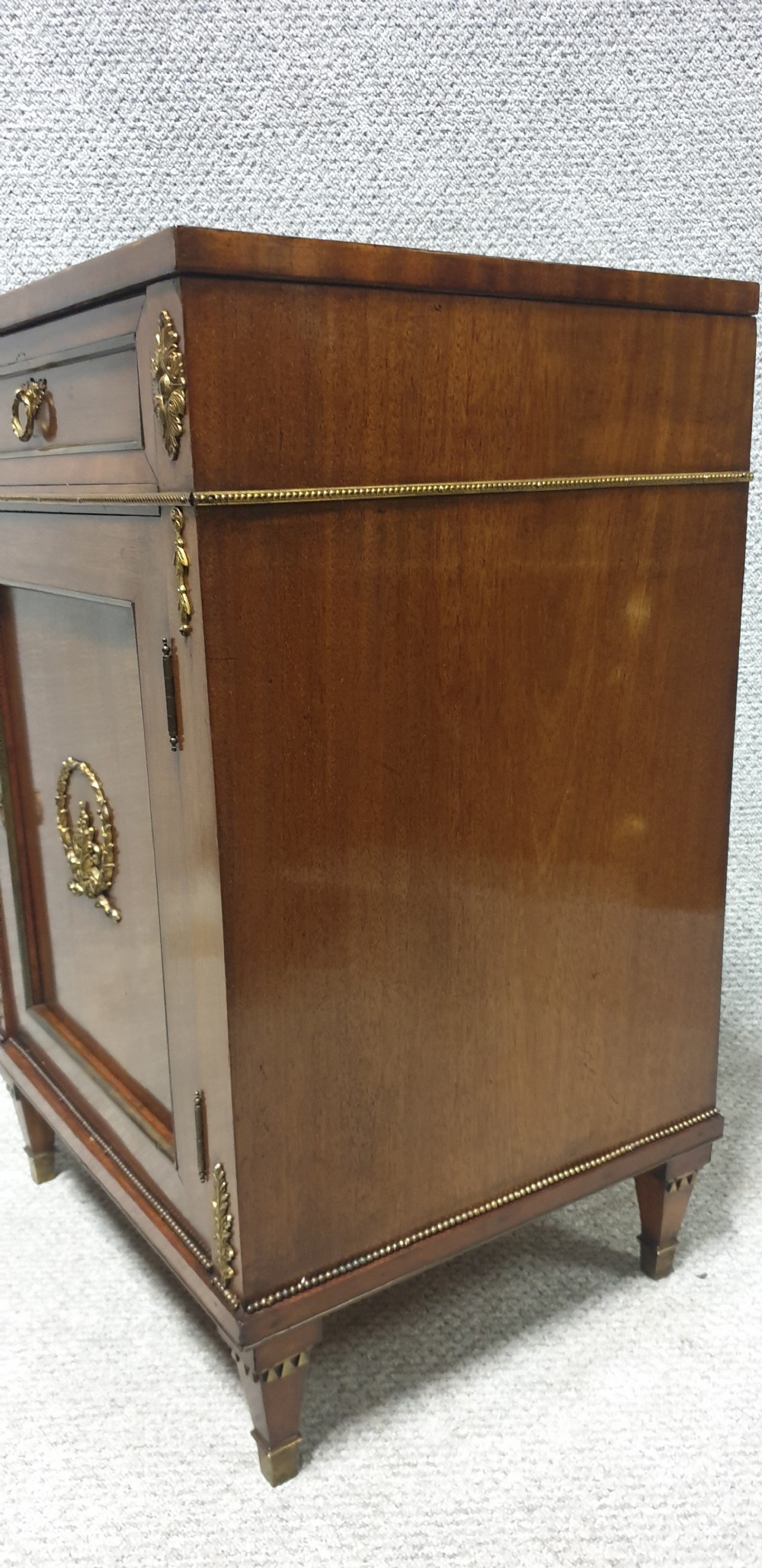 A mahogany side cabinet featuring gilt bronze mounts, a single top drawer over a cupboard door with a central laurel wreath, raised on square tapered legs. View IX