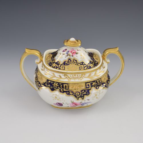 fig 1. A Regency period lidded porcelain sucrier featuring twin handles, cobalt blue borders decorated with gilt Greek key scrolling, and hand-painted floral sprays.