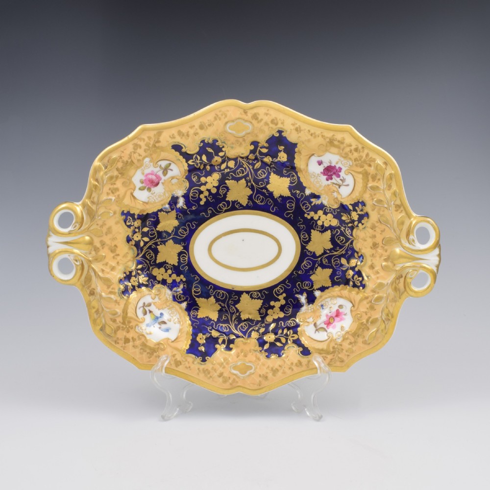 An oval antique Regency porcelain dessert dish featuring cobalt blue and gold scrollwork, hand-painted floral panels, and pierced handles.