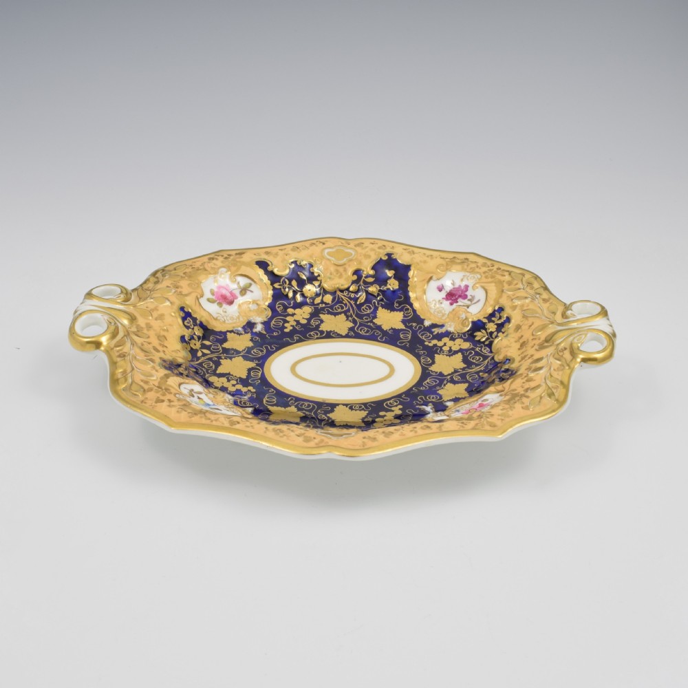 An oval antique Regency porcelain dessert dish featuring cobalt blue and gold scrollwork, hand-painted floral panels, and pierced handles. View II