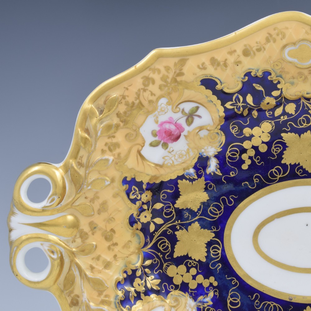 An oval antique Regency porcelain dessert dish featuring cobalt blue and gold scrollwork, hand-painted floral panels, and pierced handles. View III