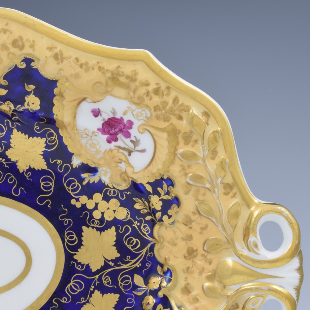 An oval antique Regency porcelain dessert dish featuring cobalt blue and gold scrollwork, hand-painted floral panels, and pierced handles. View IV