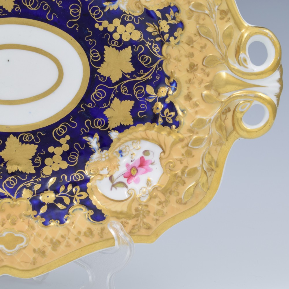 An oval antique Regency porcelain dessert dish featuring cobalt blue and gold scrollwork, hand-painted floral panels, and pierced handles. View V