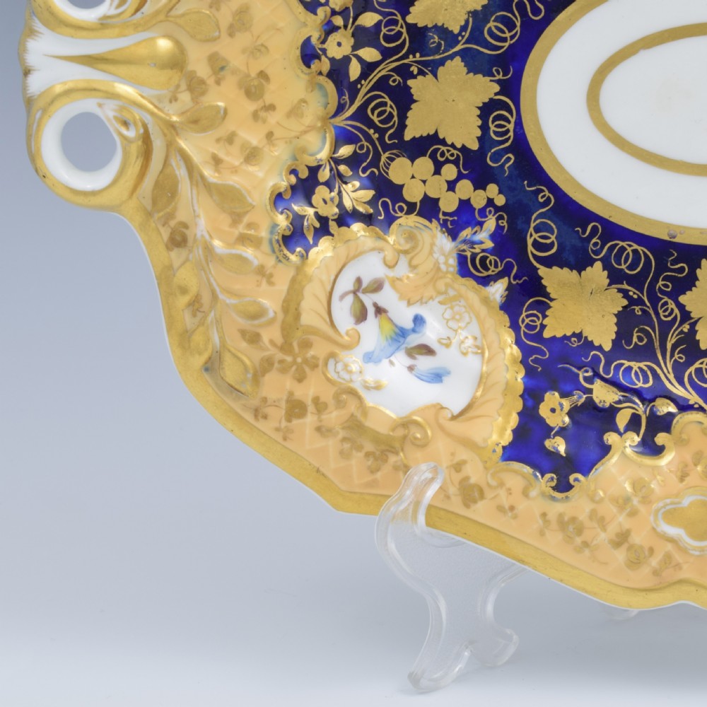 An oval antique Regency porcelain dessert dish featuring cobalt blue and gold scrollwork, hand-painted floral panels, and pierced handles. View VI