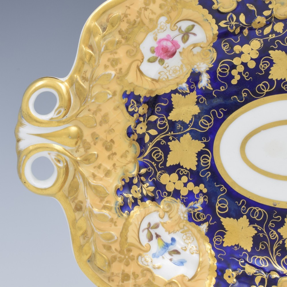 An oval antique Regency porcelain dessert dish featuring cobalt blue and gold scrollwork, hand-painted floral panels, and pierced handles. View VII