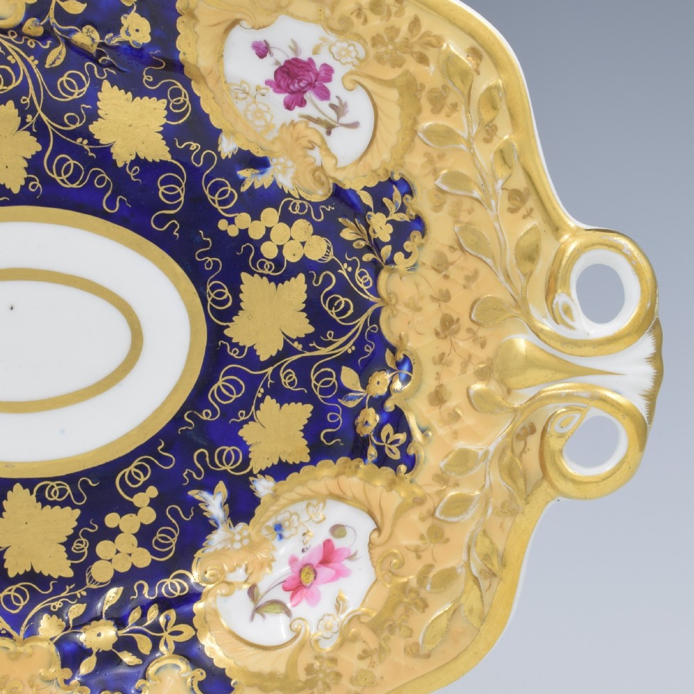 An oval antique Regency porcelain dessert dish featuring cobalt blue and gold scrollwork, hand-painted floral panels, and pierced handles. View VIII