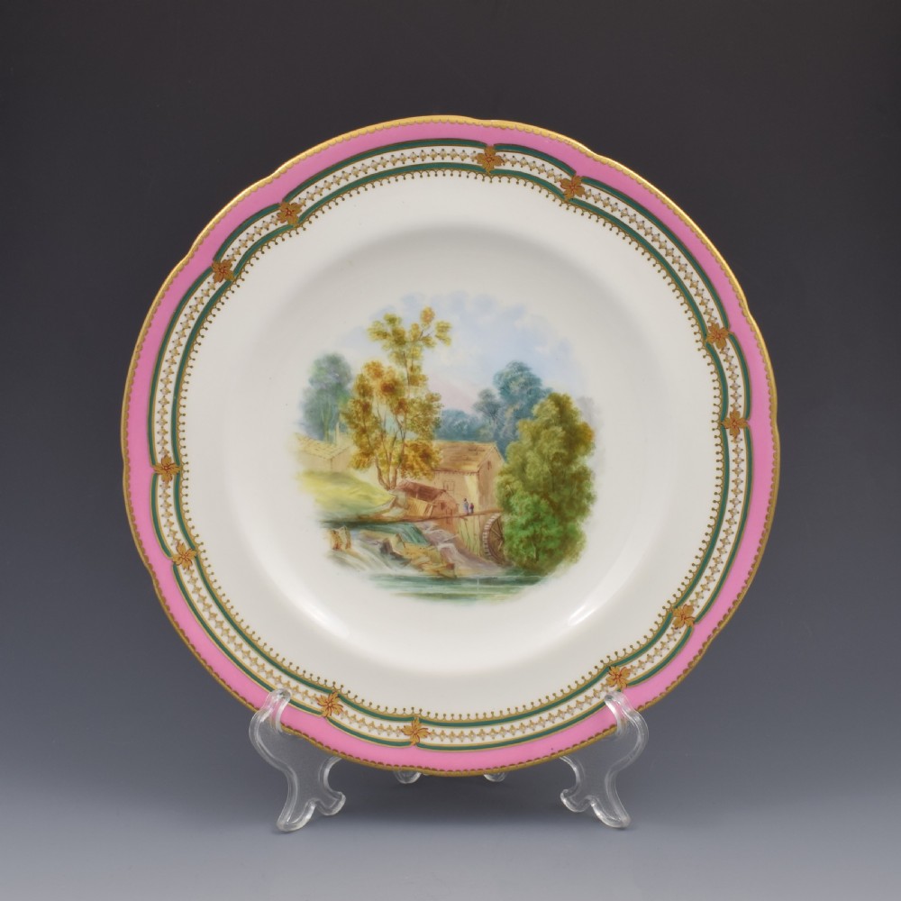 A circular porcelain cabinet plate featuring a hand-painted countryside landscape in the centre, framed by a pink ground rim with gilt and green jewelled details.