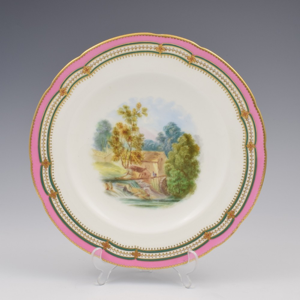 A circular porcelain cabinet plate featuring a hand-painted countryside landscape in the centre, framed by a pink ground rim with gilt and green jewelled details. View II