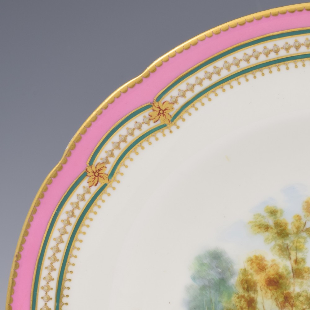 A circular porcelain cabinet plate featuring a hand-painted countryside landscape in the centre, framed by a pink ground rim with gilt and green jewelled details. View IV