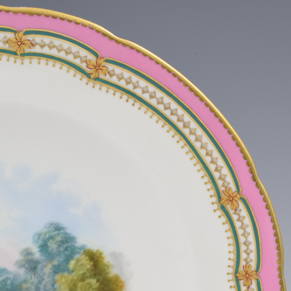 A circular porcelain cabinet plate featuring a hand-painted countryside landscape in the centre, framed by a pink ground rim with gilt and green jewelled details. View V