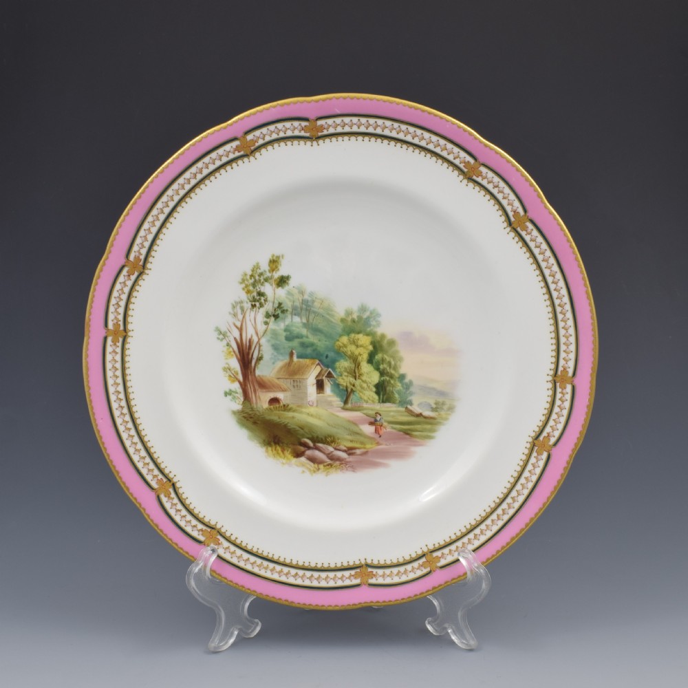 An antique porcelain dessert plate featuring a hand-painted pastoral landscape central vignette encircled by a pink border with gilded decoration and raised white enamel dots.