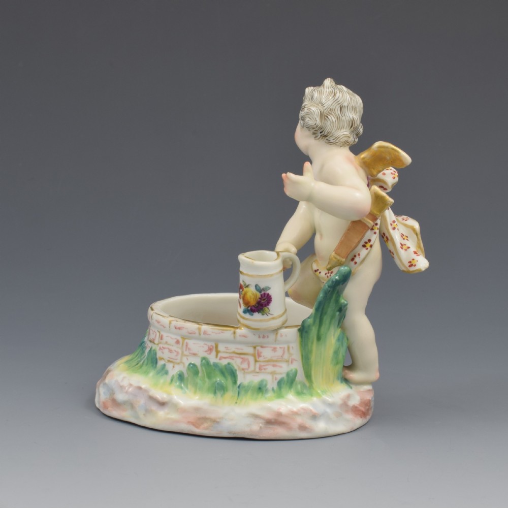 A 19th-century KPM Berlin porcelain table salt featuring a cupid figure standing beside a brick-patterned well basin with hand-painted details. View V