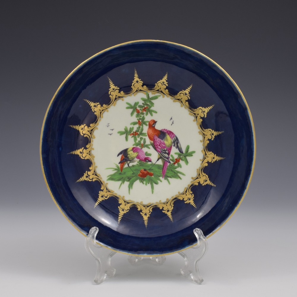 An eighteenth-century circular porcelain saucer dish featuring a deep cobalt blue border with gilt scrollwork and a white central medallion containing hand-painted exotic birds.