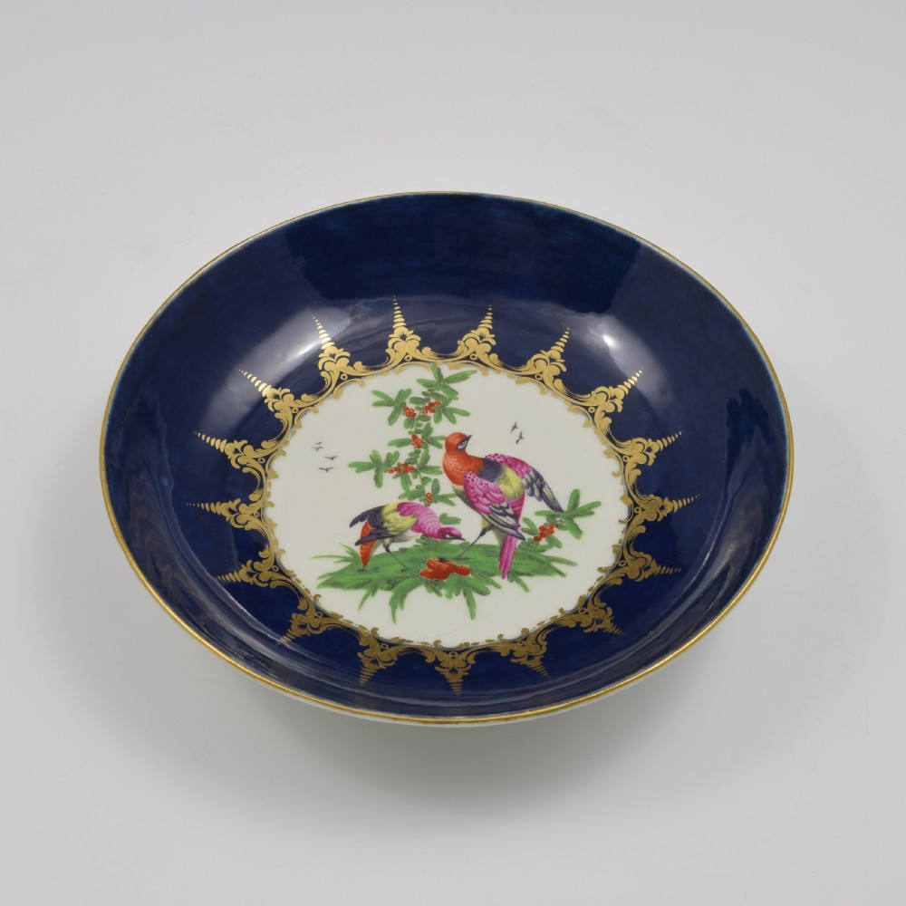 An eighteenth-century circular porcelain saucer dish featuring a deep cobalt blue border with gilt scrollwork and a white central medallion containing hand-painted exotic birds. View V