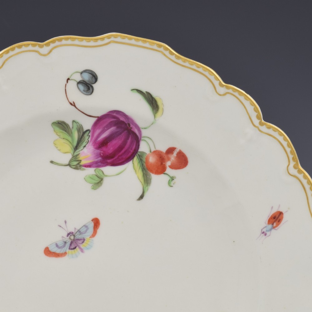 An eighteenth-century soft-paste porcelain dessert plate with a lobed gilded rim, decorated with hand-painted fruit motifs and insects against a white background. View II