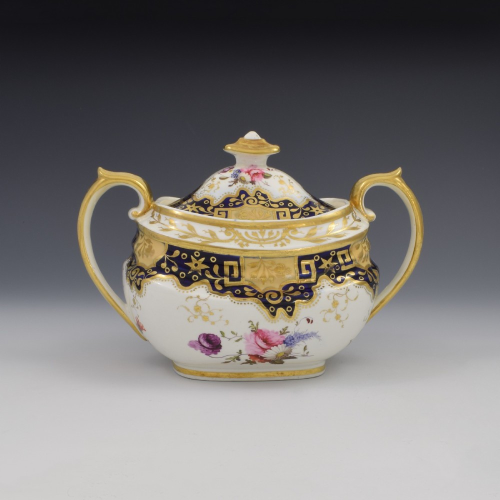A Regency period lidded porcelain sucrier featuring twin handles, cobalt blue borders decorated with gilt Greek key scrolling, and hand-painted floral sprays.