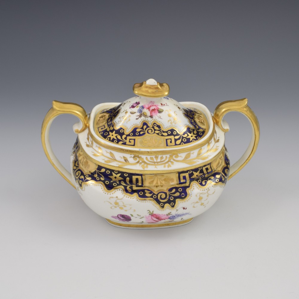 A Regency period lidded porcelain sucrier featuring twin handles, cobalt blue borders decorated with gilt Greek key scrolling, and hand-painted floral sprays. View II