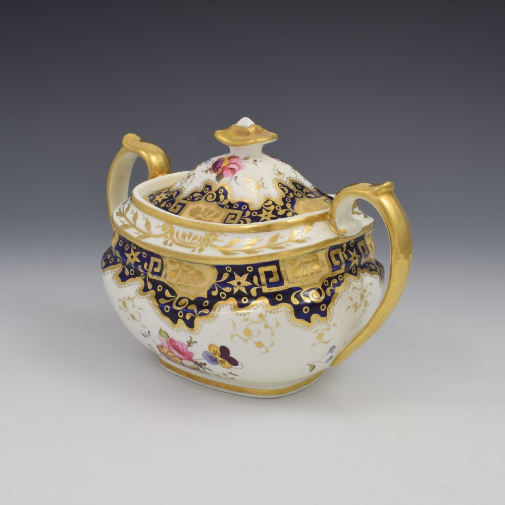 A Regency period lidded porcelain sucrier featuring twin handles, cobalt blue borders decorated with gilt Greek key scrolling, and hand-painted floral sprays. View V