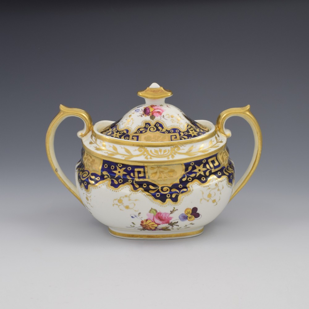 A Regency period lidded porcelain sucrier featuring twin handles, cobalt blue borders decorated with gilt Greek key scrolling, and hand-painted floral sprays. View VI