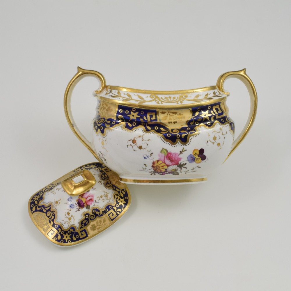 A Regency period lidded porcelain sucrier featuring twin handles, cobalt blue borders decorated with gilt Greek key scrolling, and hand-painted floral sprays. View XII