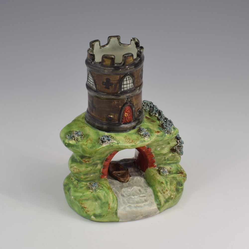 An antique Staffordshire pearlware spill vase shaped as a brown castellated turret standing over a green rocky bridge with a small boat underneath.