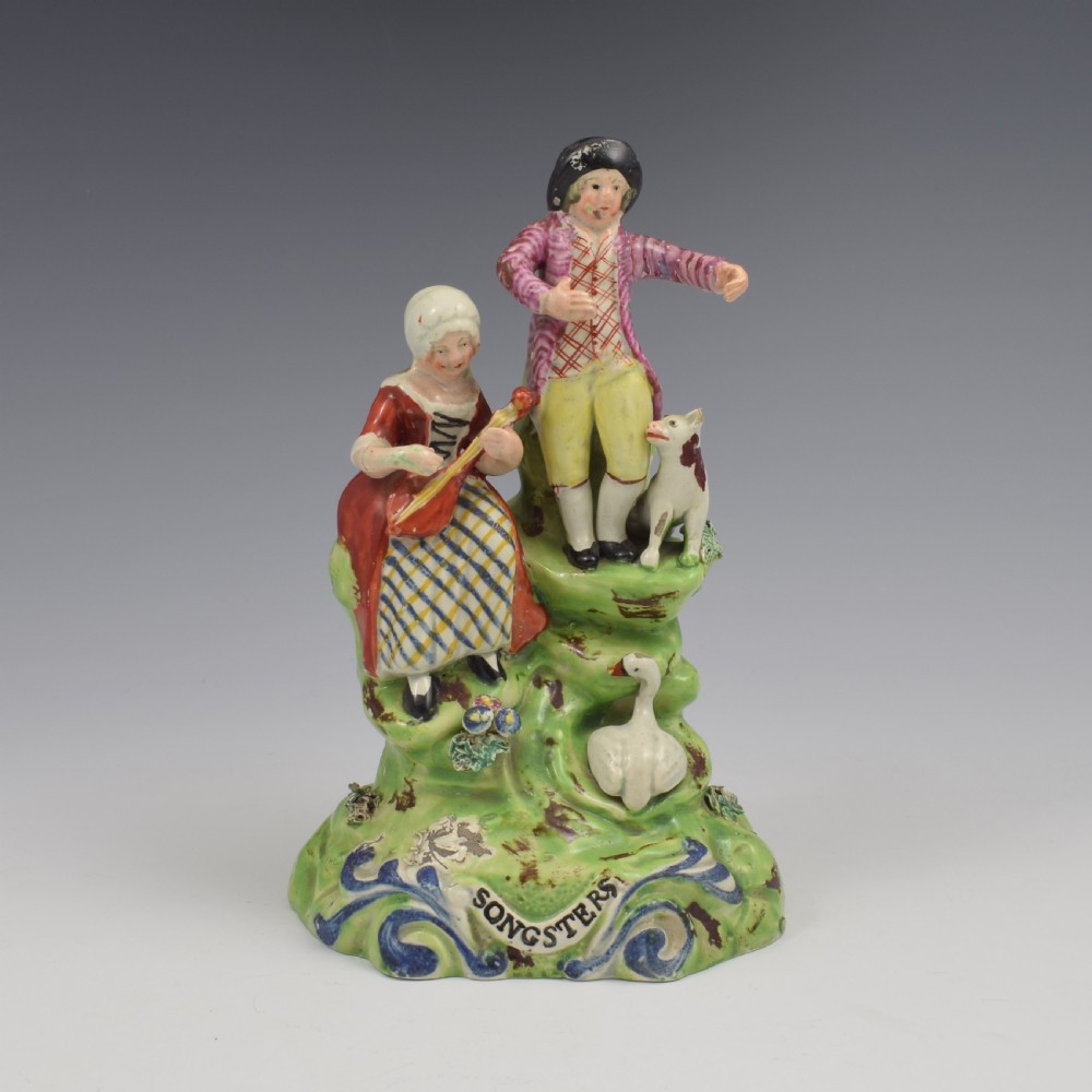 An antique Staffordshire pearlware figure group titled Songsters depicting a seated woman playing an instrument alongside a standing man, a dog, and a swan on a green mound base.