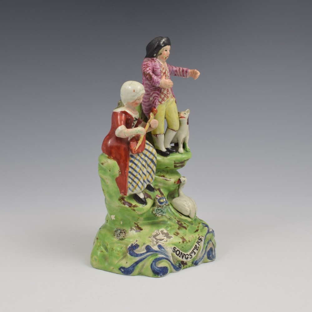 An antique Staffordshire pearlware figure group titled Songsters depicting a seated woman playing an instrument alongside a standing man, a dog, and a swan on a green mound base. View II