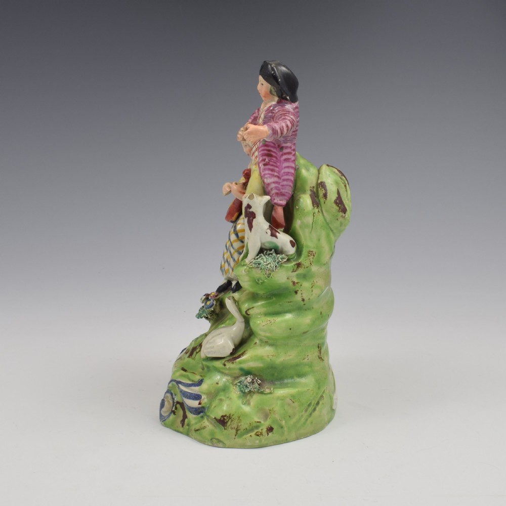 An antique Staffordshire pearlware figure group titled Songsters depicting a seated woman playing an instrument alongside a standing man, a dog, and a swan on a green mound base. View VI
