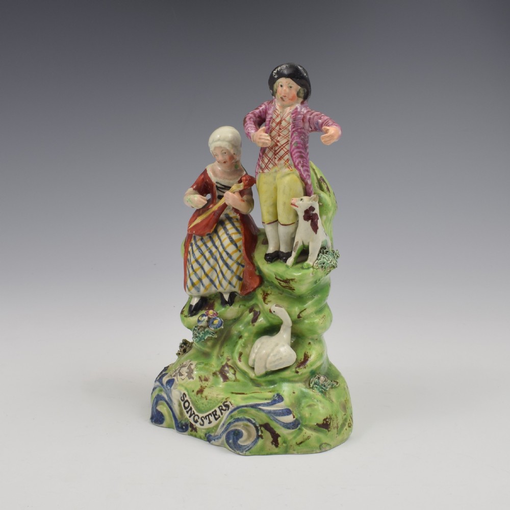 An antique Staffordshire pearlware figure group titled Songsters depicting a seated woman playing an instrument alongside a standing man, a dog, and a swan on a green mound base. View VII