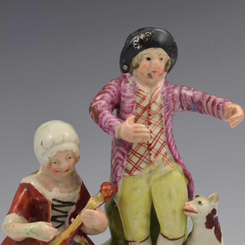 An antique Staffordshire pearlware figure group titled Songsters depicting a seated woman playing an instrument alongside a standing man, a dog, and a swan on a green mound base. View IX