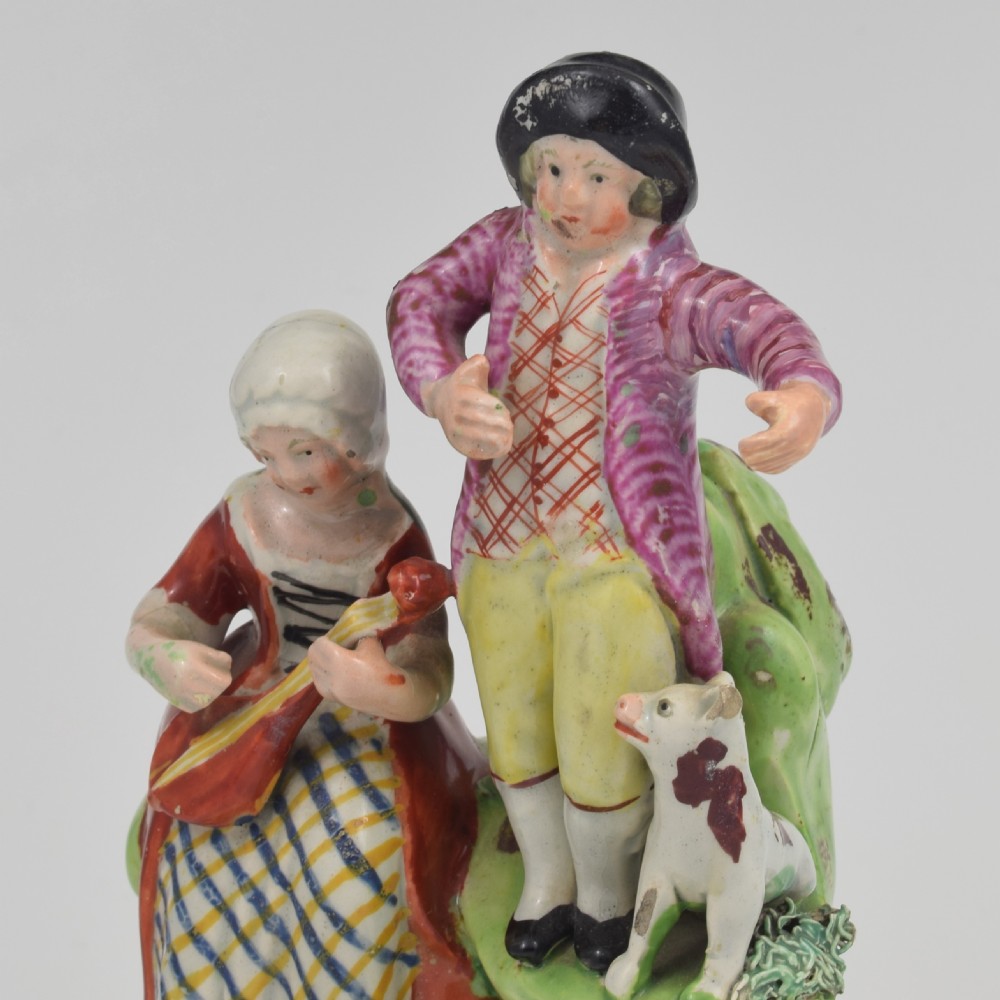 An antique Staffordshire pearlware figure group titled Songsters depicting a seated woman playing an instrument alongside a standing man, a dog, and a swan on a green mound base. View XII