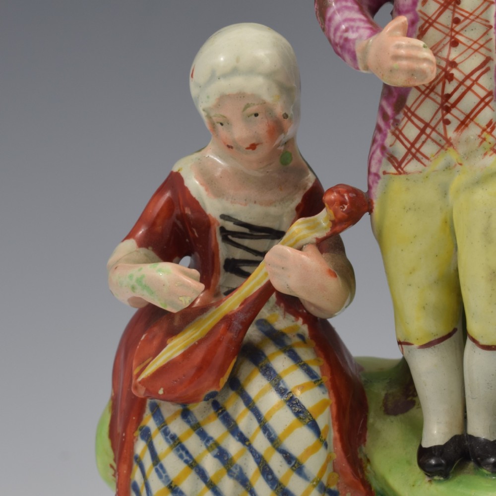 An antique Staffordshire pearlware figure group titled Songsters depicting a seated woman playing an instrument alongside a standing man, a dog, and a swan on a green mound base. View XV