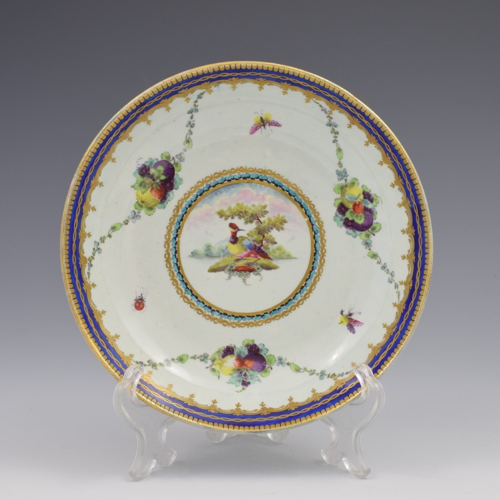 A circular eighteenth-century Worcester porcelain saucer dish decorated with a hand-painted bird in a central medallion, fruit garlands, and a gilded cobalt blue border.