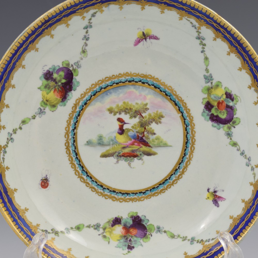 A circular eighteenth-century Worcester porcelain saucer dish decorated with a hand-painted bird in a central medallion, fruit garlands, and a gilded cobalt blue border. View II