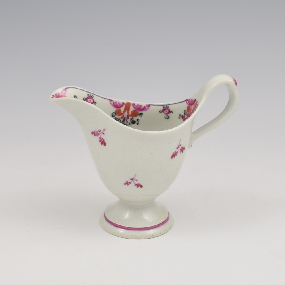 An 18th-century Georgian New Hall white porcelain cream jug featuring a high clip handle, pedestal foot, and hand-painted pink floral motifs.