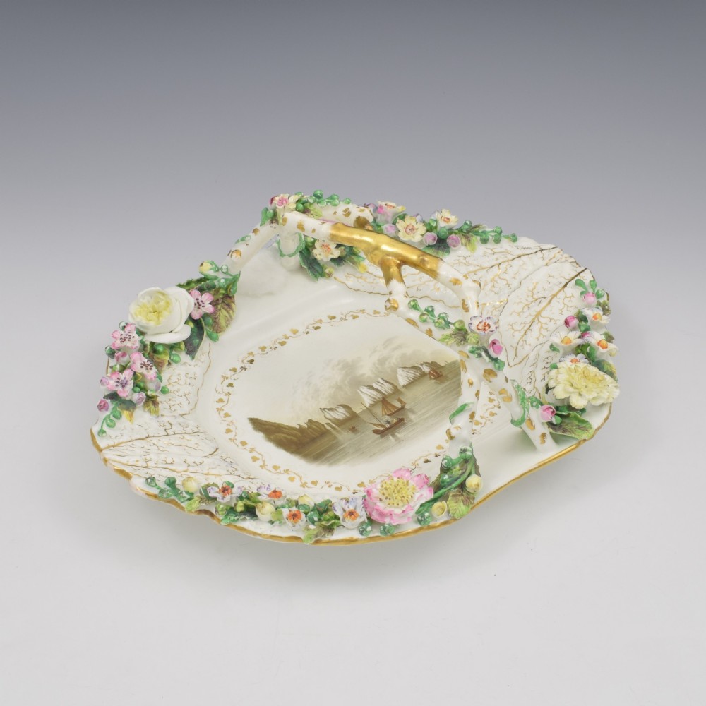 A white porcelain basket decorated with hand-sculpted flowers along the rim, a gilded twig-shaped handle, and a hand-painted marine landscape scene in the centre.