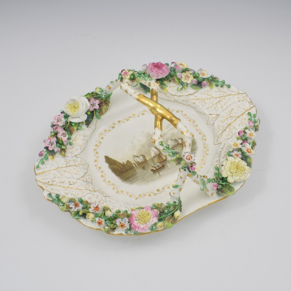 A white porcelain basket decorated with hand-sculpted flowers along the rim, a gilded twig-shaped handle, and a hand-painted marine landscape scene in the centre. View II