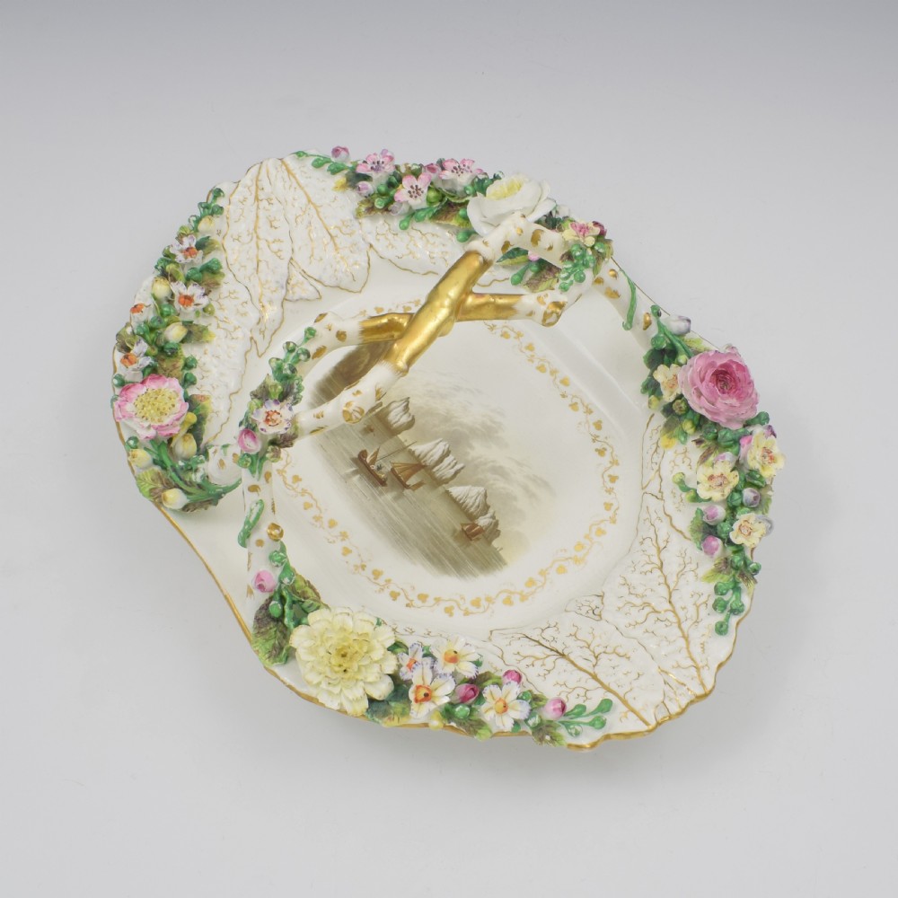 A white porcelain basket decorated with hand-sculpted flowers along the rim, a gilded twig-shaped handle, and a hand-painted marine landscape scene in the centre. View IV