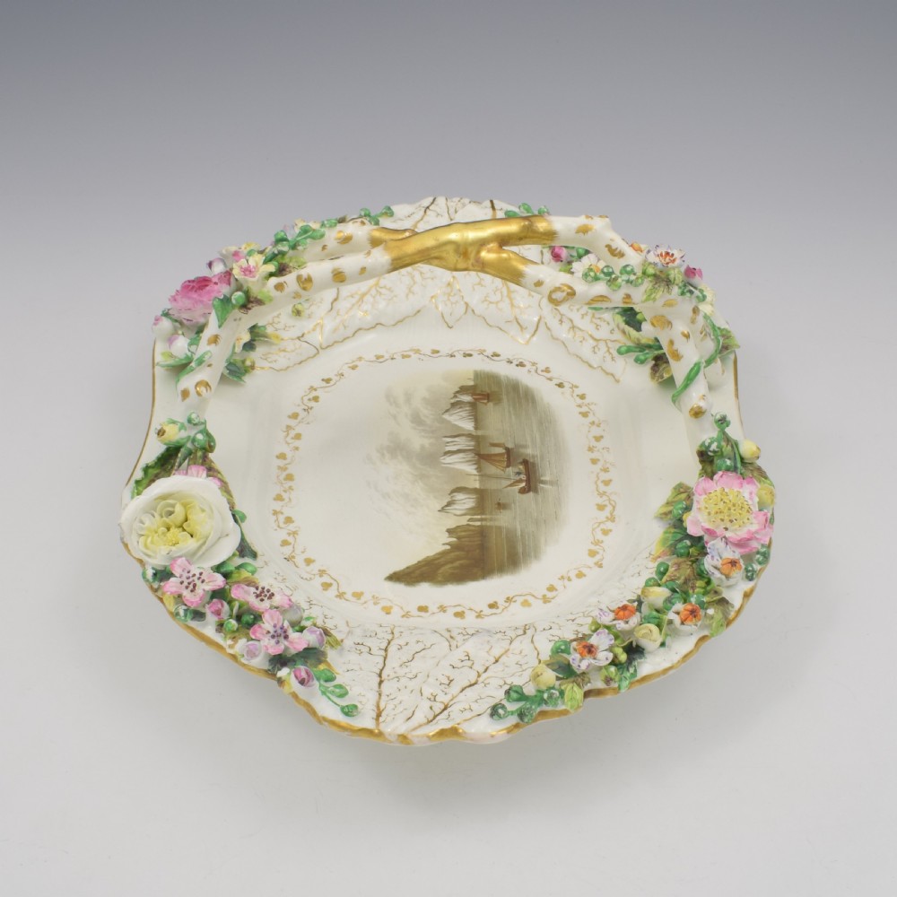 A white porcelain basket decorated with hand-sculpted flowers along the rim, a gilded twig-shaped handle, and a hand-painted marine landscape scene in the centre. View V